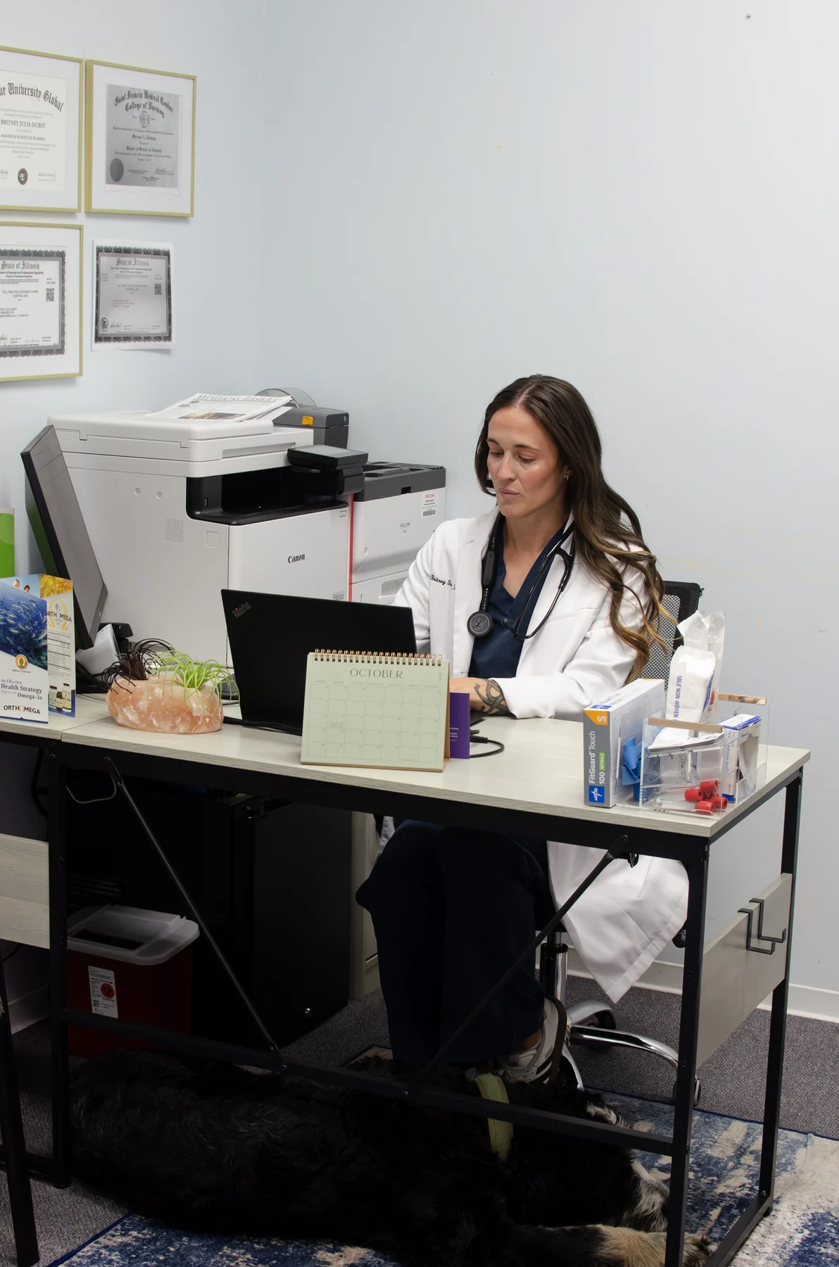 Clinician in a white coat and stethoscope working at an office laptop